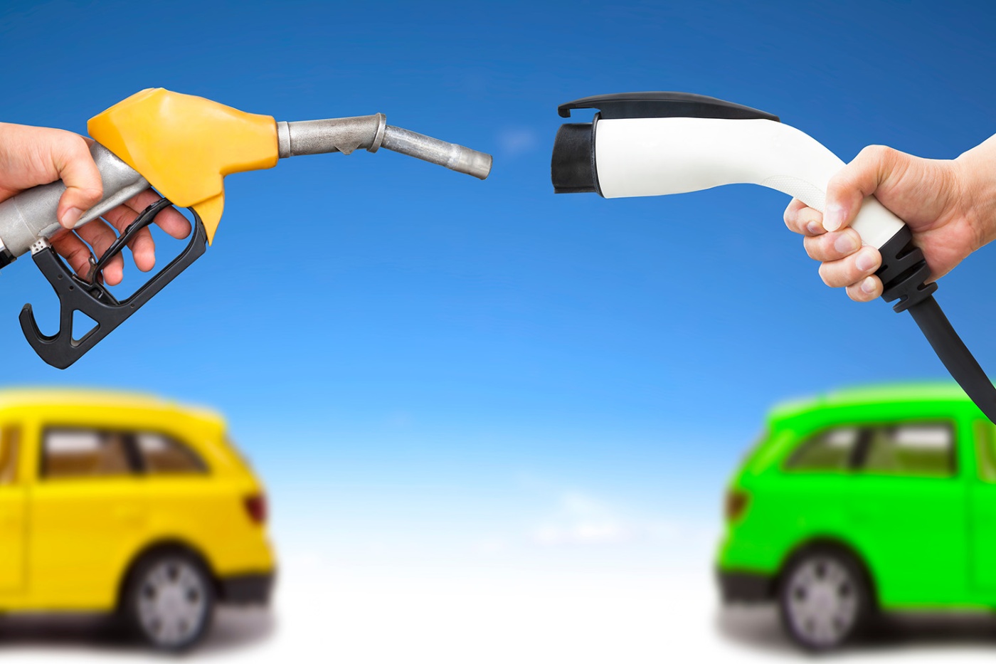 One hand holds a yellow fuel nozzle, another hand holds a white electric charging plug, with two small cars, yellow and green, in the background against a blue sky.