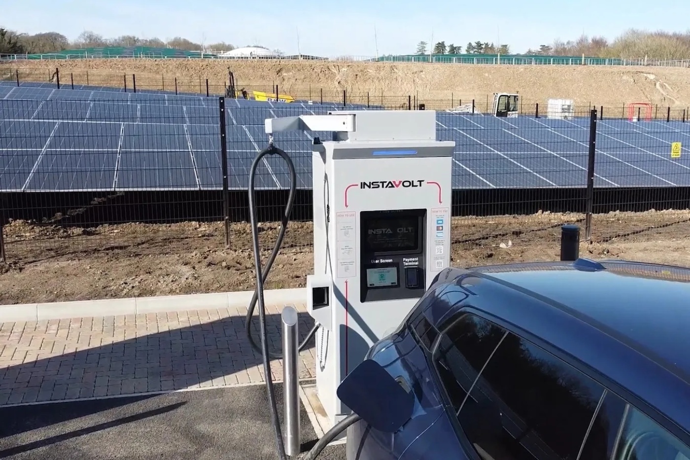 Electric car charging at a white Instavolt charging station in front of a large solar panel field.