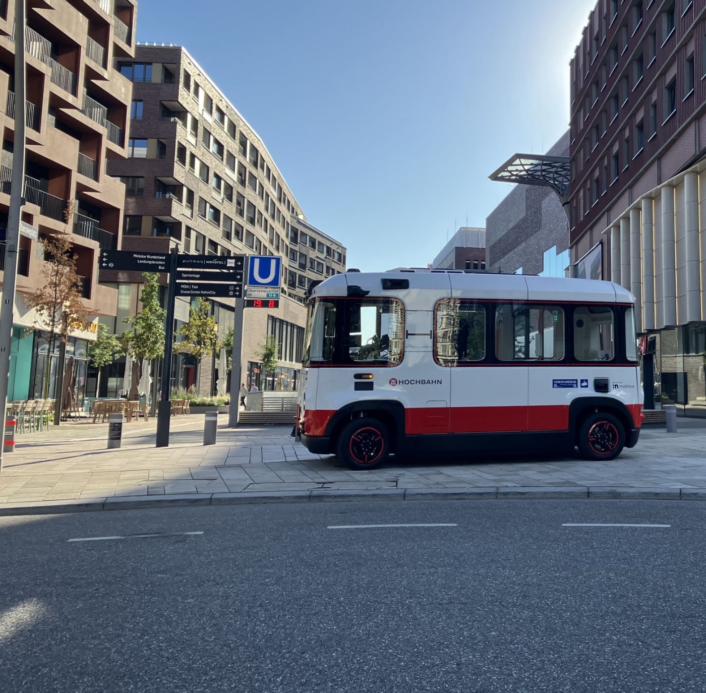 Small red and white bus with 'HOCHBAHN' lettering parked on a street in a modern urban area with multi-story buildings and clear sky.
