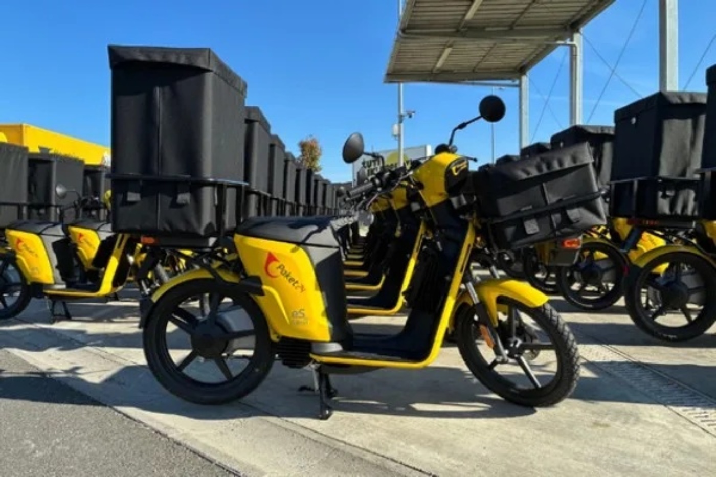 Yellow postal motor scooter with black cargo box parked among multiple similar vehicles.