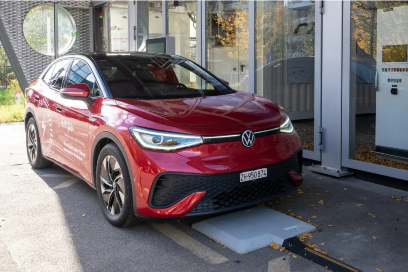 Ein rotes Elektroauto steht über einer induktiven Ladestation vor einem modernen Gebäude.