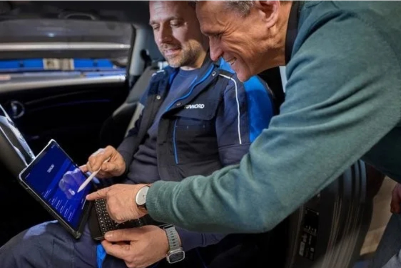Zwei Männer im Auto betrachten Diagnosedaten auf einem Tablet, einer zeigt mit einem Stift darauf.