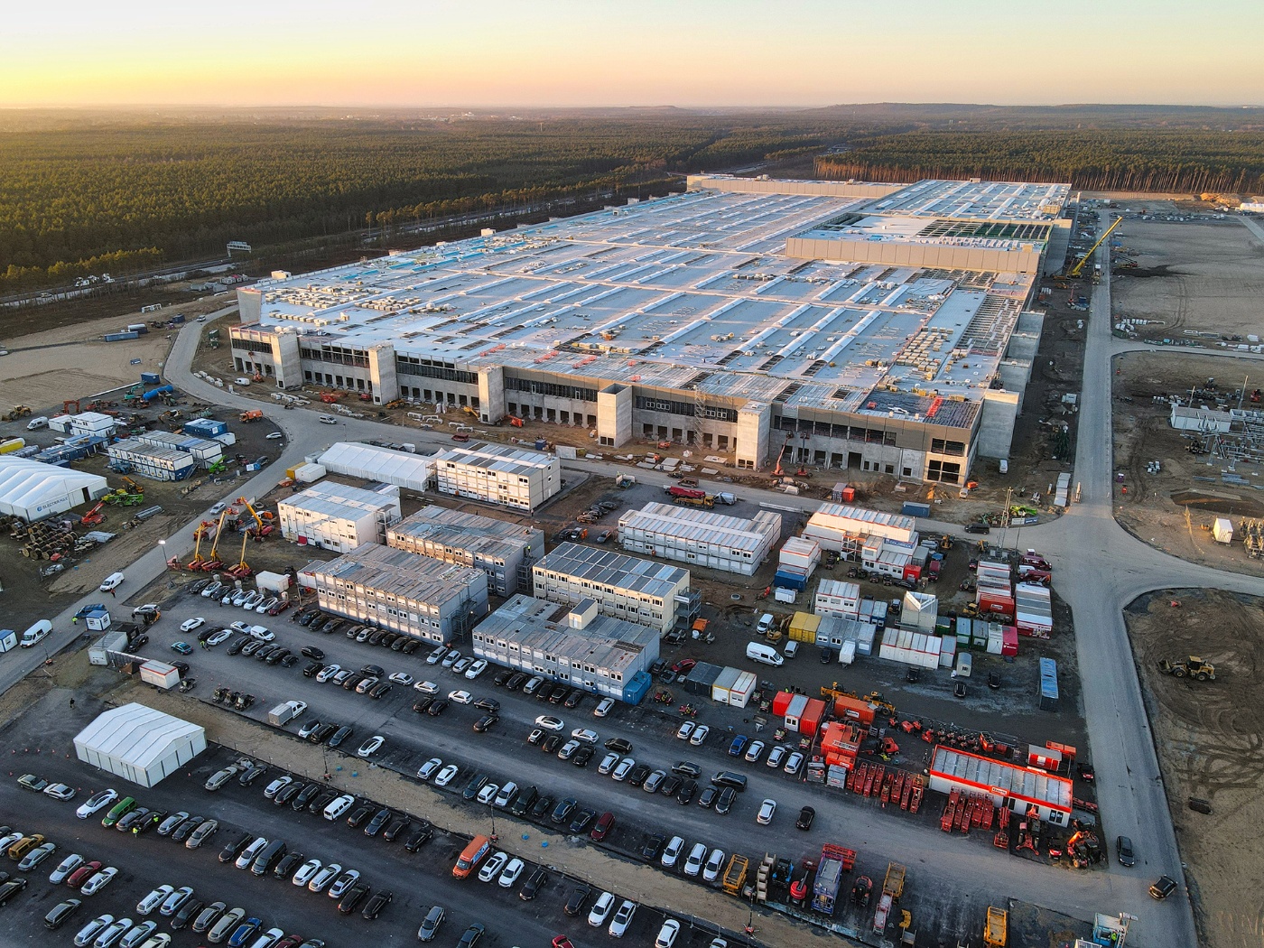 Luftaufnahme einer im Bau befindlichen Tesla-Fabrik mit parkenden Autos und Wald im Hintergrund.