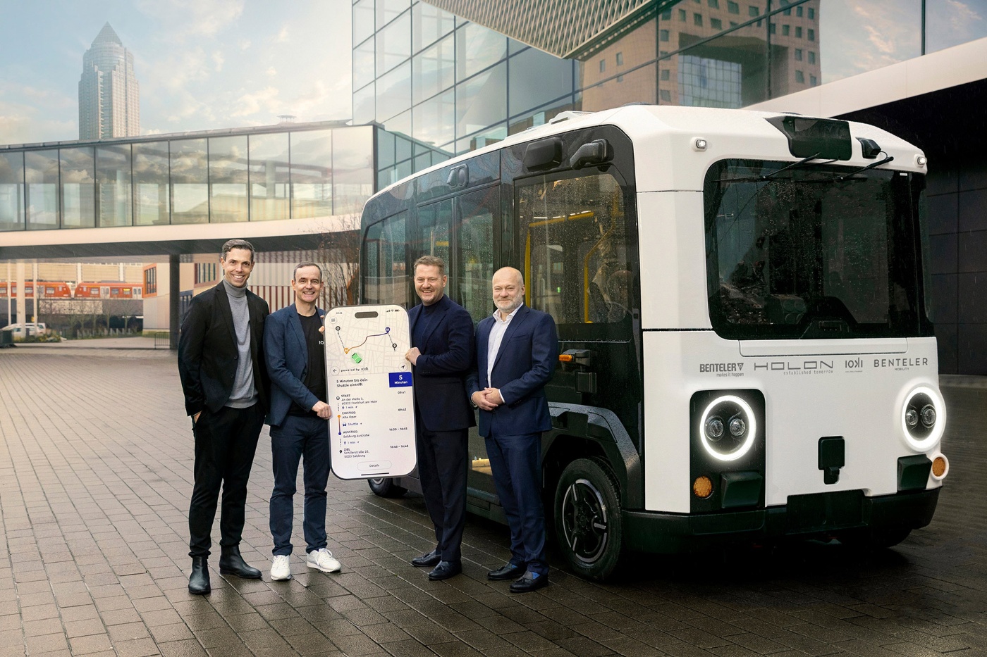 Vier Männer stehen neben einem autonomen Shuttle und halten ein großes Smartphone-Schild.
