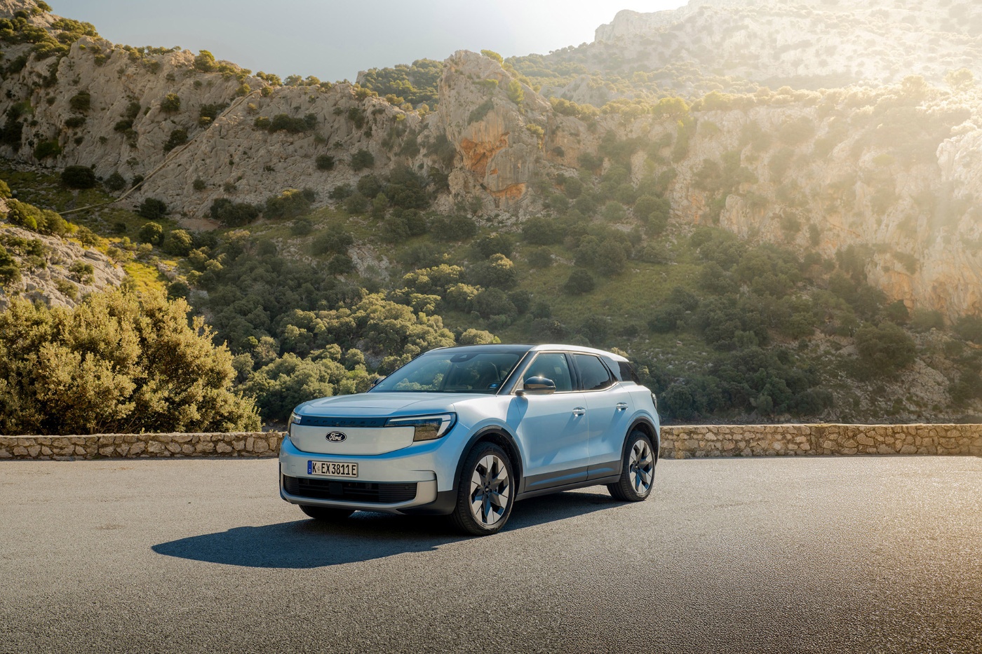 A light blue electric SUV from Ford is parked on a mountain road in front of rocky hills.