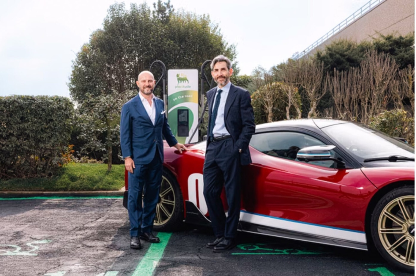 Zwei Männer im Anzug stehen neben einem roten Elektro-Sportwagen an einer Ladesäule.