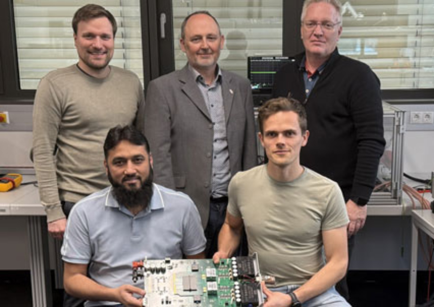 Five men in lab standing and sitting with a circuit board.