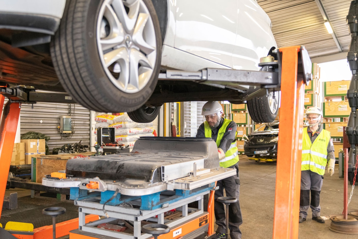 Worker in factory handling EV battery module under vehicle lift.