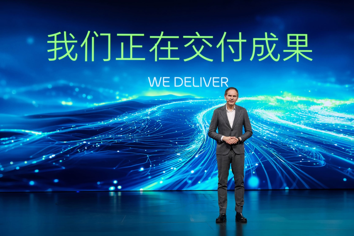 Moderator auf Bühne vor blauem Digitalhintergrund mit Text „WE DELIVER“.