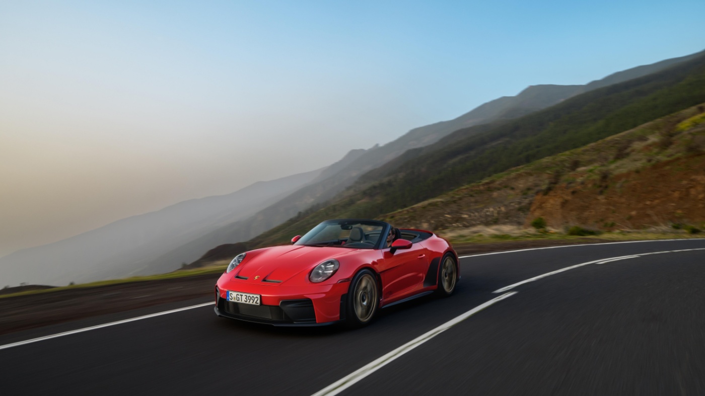 Roter Porsche-Sportwagen fährt auf Küstenstraße mit Hügeln bei Sonnenuntergang.