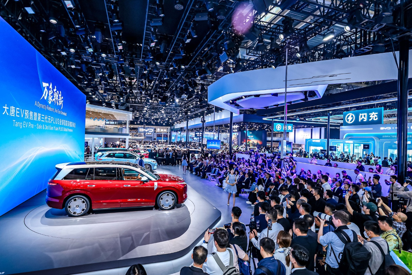 Red car on display at a crowded auto show with large screen.