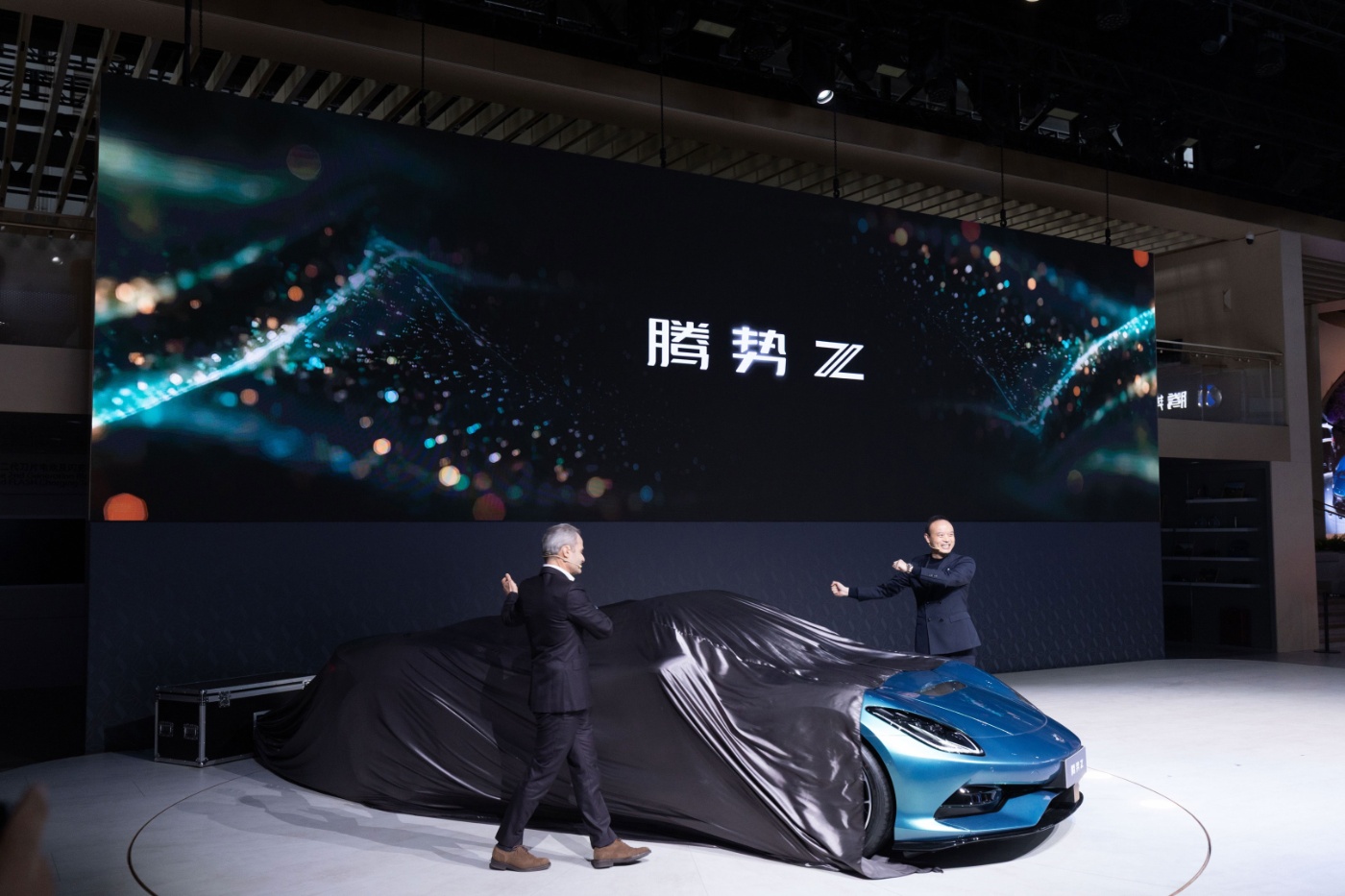Two people unveil a blue car on a stage with large screen behind.
