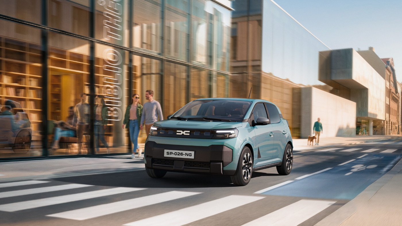 Türkisfarbener Dacia-Kleinwagen fährt durch eine moderne Innenstadt mit Glasfassaden, Fußgängern und einem Zebrastreifen.