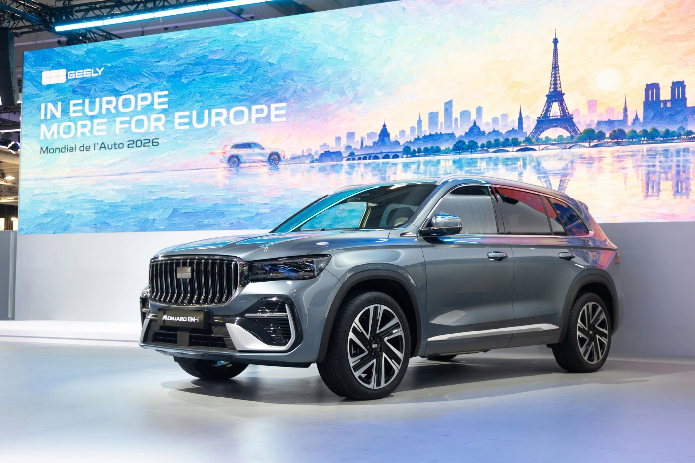Grauer Geely SUV auf einer Automesse vor einer Kulisse mit europäischen Wahrzeichen.