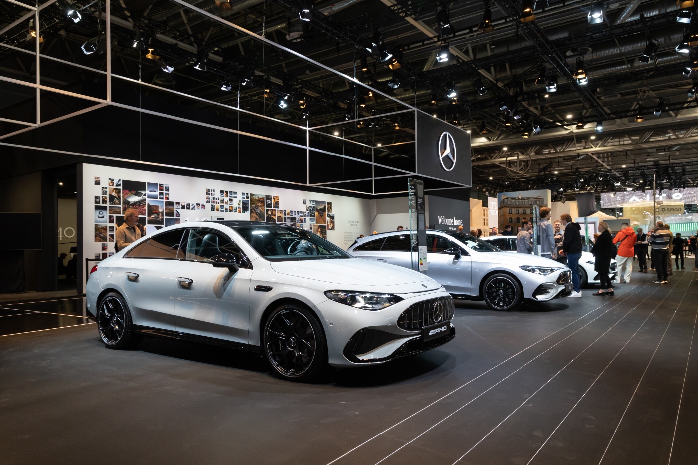 Weißer Mercedes-AMG mit sportlicher Limousinenkarosserie und schwarzem Kühlergrill auf einem beleuchteten Mercedes-Benz-Messestand mit weiteren Fahrzeugen und Besuchern.