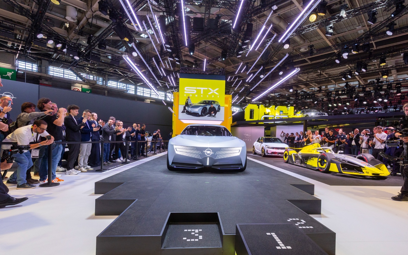 Opel präsentiert das STX-Konzeptauto auf einer Automesse, umgeben von Besuchern und Rennwagen.